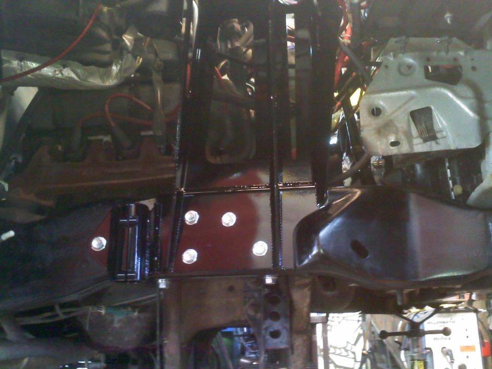 Close-up of a vehicle's frame with black painted suspension parts and silver bolts. Watermark reads SUPERMOTORS.NET