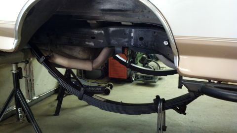 1996 Ford Bronco - A tall multi-leaf spring pack sits on the shop floor in front of a tan-and-white body panel raised on jack stands, with the empty wheel arc…