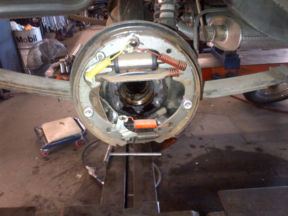 Close-up of a car's rear brake drum assembly with brake shoes and springs visible. A Mobil oil drum is in the background