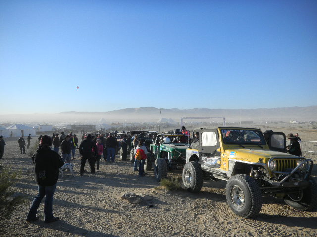 Off-road vehicles lined up on a dirt lot with people standing around, mountains in the background
