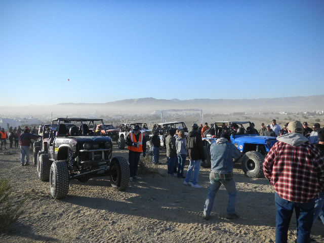 Group of off-road vehicles and people gathered on a dirt lot with hills in the background