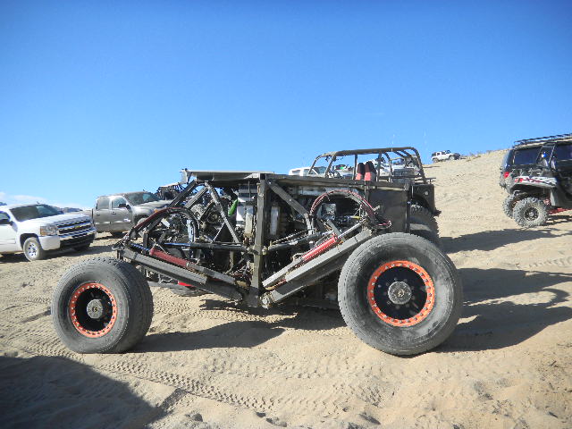 Custom off-road buggy with exposed engine and roll cage, parked on sand with other vehicles in background