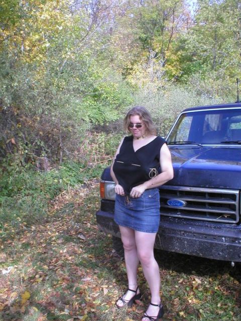 1989 Ford Bronco - A woman in sunglasses, a black top and denim skirt poses beside the front end of a dirt-splattered blue square-body Ford truck parked on a…