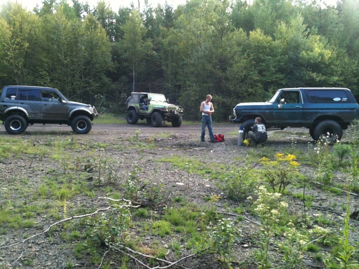 Blue lifted Ford Bronco with two other 4x4s parked in a wooded gravel clearing