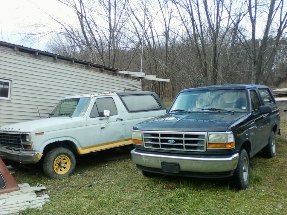 White 1980s Ford Bronco with yellow wheel parked next to a dark green later-model Bronco on grass by a building