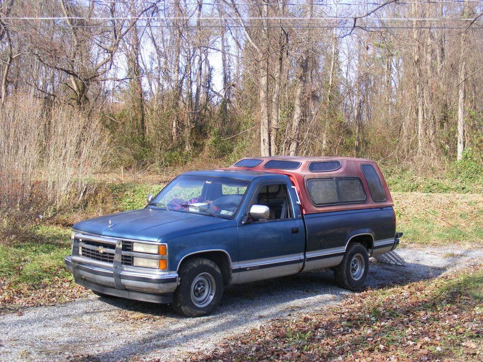 Blue Chevrolet C/K pickup with red camper shell parked on gravel near bare trees