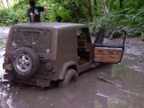 1987 Mazda RX-7 - A muddy Jeep Wrangler is stuck in a ditch, with two people standing nearby in a wooded area.