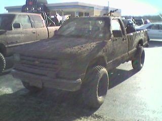 1987 Mazda RX-7 - A heavily mud-caked pickup truck sits parked, with another vehicle partially visible to its left.