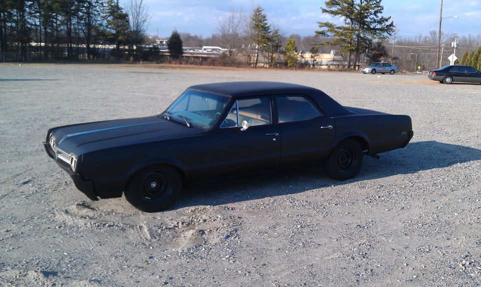 Black 1966 Oldsmobile Cutlass sedan parked on gravel lot, matte finish, black wheels