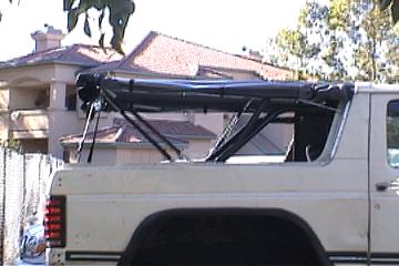 1991 Ford Bronco - The rear section of an SUV shows a black soft top frame, with a building in the background.