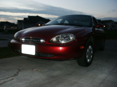 1998 Mercury Sable - The front end of a red sedan is visible parked in front of houses under a cloudy sky.
