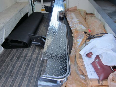 1987 Dodge 1500 - A diamond plate rear bumper with chrome trim is stored in the bed of a vehicle, alongside other parts and documents.