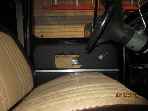 1979 Ford Bronco - The interior of a vehicle shows a tan seat and black door panel with a chrome window crank and door handle.
