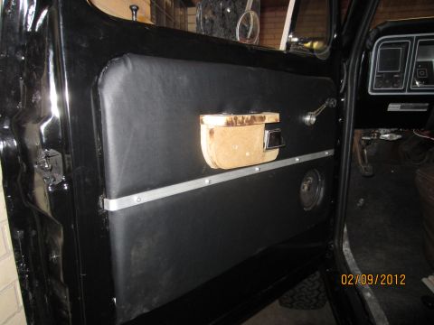 1979 Ford Bronco - Interior view of a black door panel with aftermarket wooden armrest and speaker. Dashboard gauges are visible.