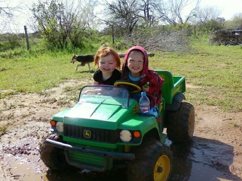 1989 Ford Bronco - Two smiling children ride in a green toy tractor, splashing through puddles in a muddy outdoor area.