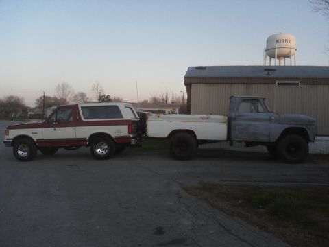 1989 Ford Bronco - A red and white Ford Bronco is parked next to a pickup truck in front of a building with a water tower.