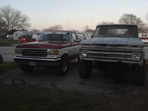 1989 Ford Bronco - A red and white 1989 Ford Bronco is parked next to an older grey truck, both appearing to be modified for off-road use.