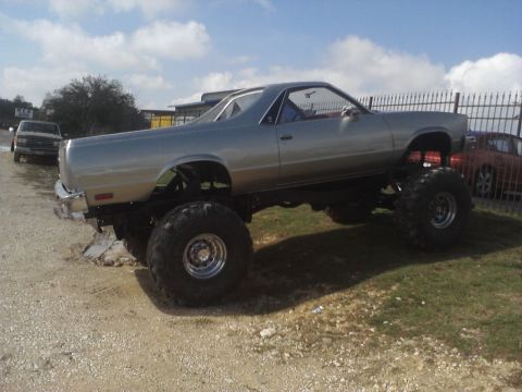 1989 Ford Bronco - A heavily modified pickup truck with oversized tires sits on a dirt lot under a partly cloudy sky.