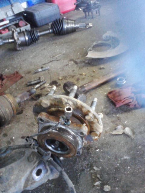 1989 Ford Bronco - Various vehicle parts, including a hub assembly and CV axle, are scattered on a concrete garage floor, suggesting a repair or disassembly p…