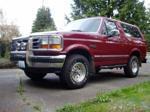 1993 Ford Bronco - A red Ford Bronco is parked on a driveway next to another vehicle, with trees in the background.