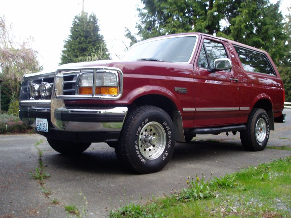 Red 1993 Ford Bronco parked next to a white truck. The Bronco has chrome wheels and a side step