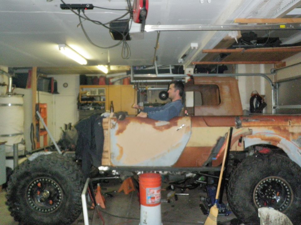 Man sits in the driver's seat of a heavily modified, rusty vehicle with large off-road tires in a garage