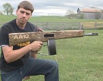 2002 Chevrolet Silverado 1500 - A man kneels in a grassy field, holding a wooden replica of an AA12 automatic shotgun. Text reading 'AA12' is visible on the replica.