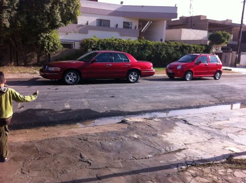 1998 Ford Crown Victoria - A red 1998 Ford Crown Victoria is parked on the street next to a smaller red car.