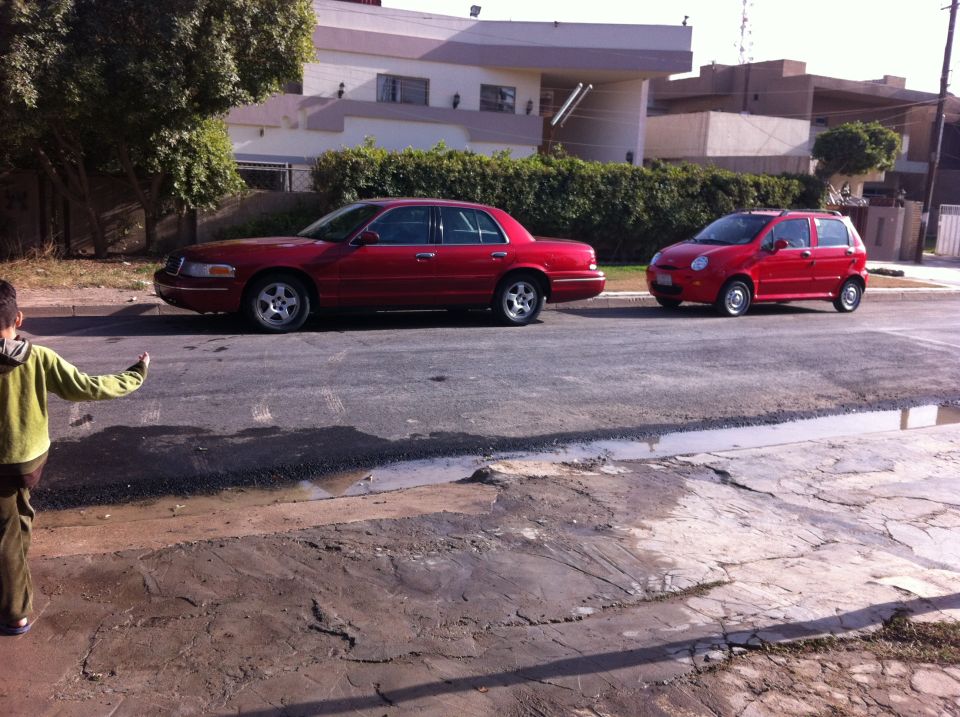 Red 1998 Ford Crown Victoria parked curbside, side view, with a red hatchback behind it
