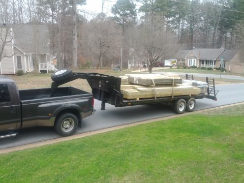 1979 Ford F-150 - A dark-colored pickup truck tows a large flatbed trailer piled high with lumber down a residential street.