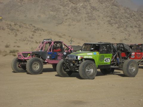 1996 Ford Bronco - Two custom off-road vehicles are parked side-by-side in a dusty, desert environment.