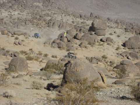 1996 Ford Bronco - Off-road vehicles traverse a dusty, boulder-strewn desert terrain, with a person observing from a rocky outcrop.