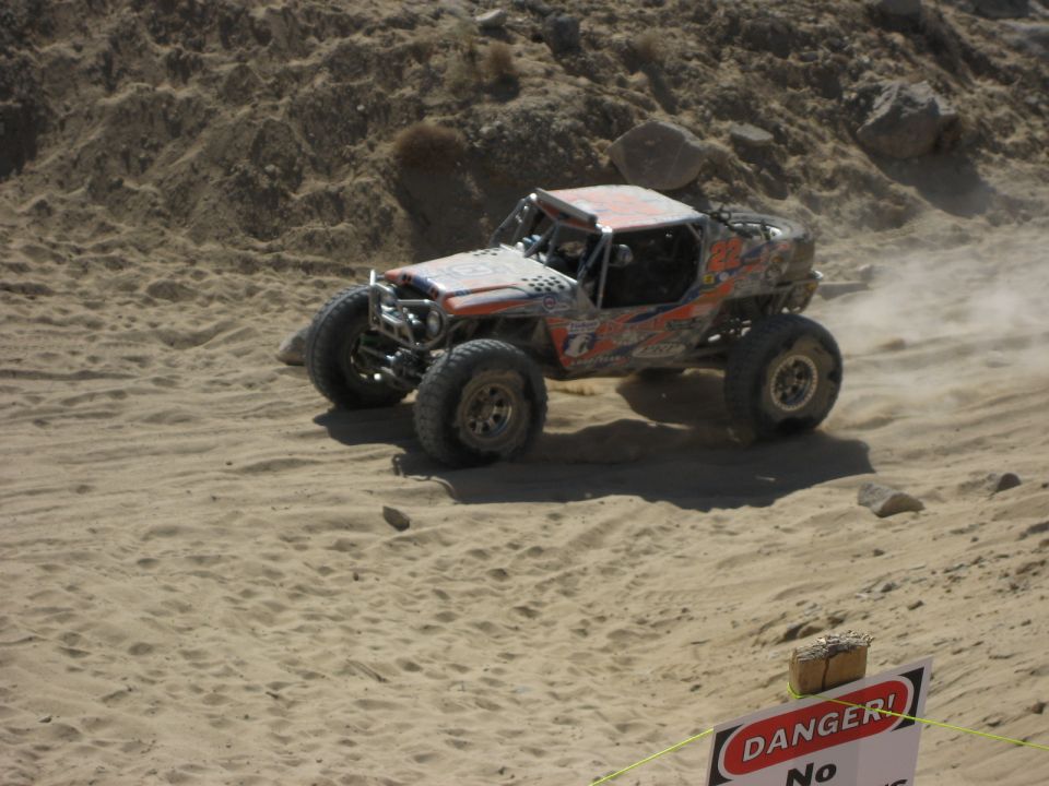 Off-road buggy with orange and black graphics kicking up dust on a sandy hill. Supermotors.net logo visible