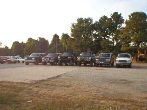 1989 Ford Bronco - A lineup of trucks and cars are parked in a dusty lot under a bright sky.