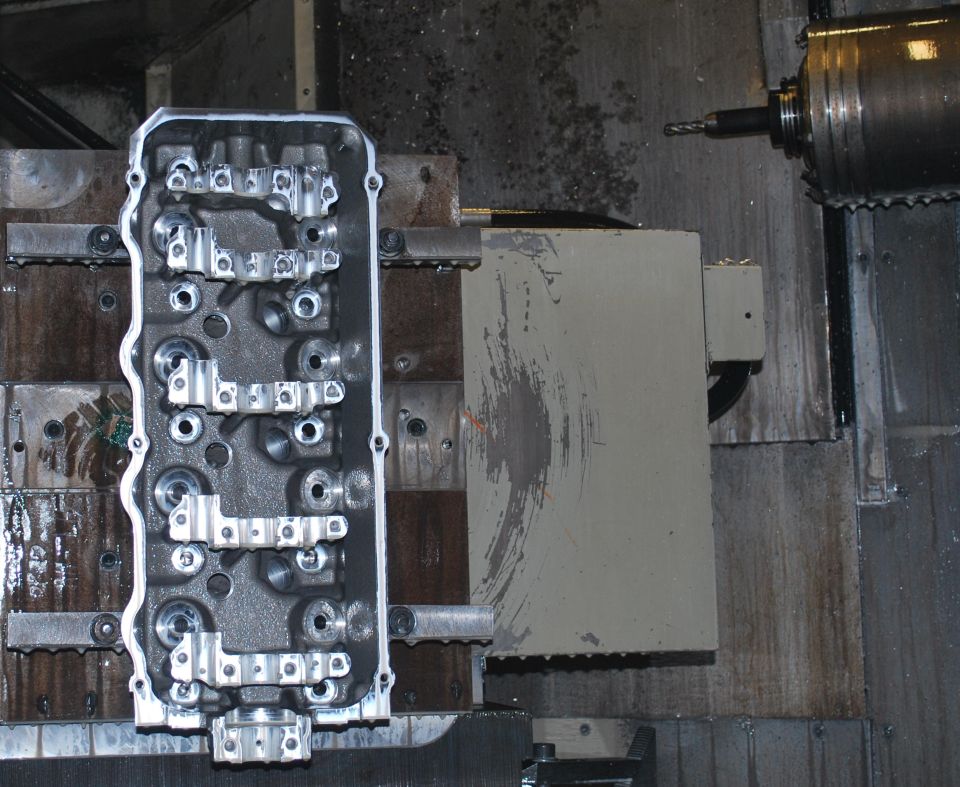 Engine cylinder head on a machining table with a CNC mill in the background