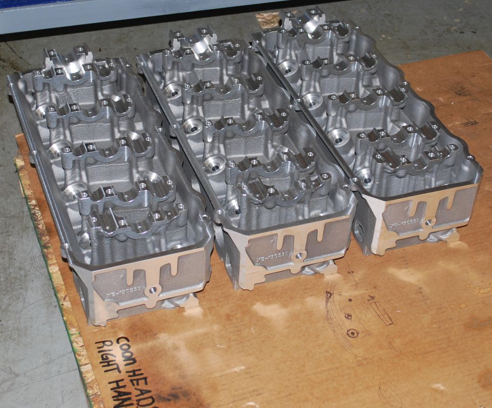 Three aluminum engine cylinder heads on a wooden surface with handwritten text