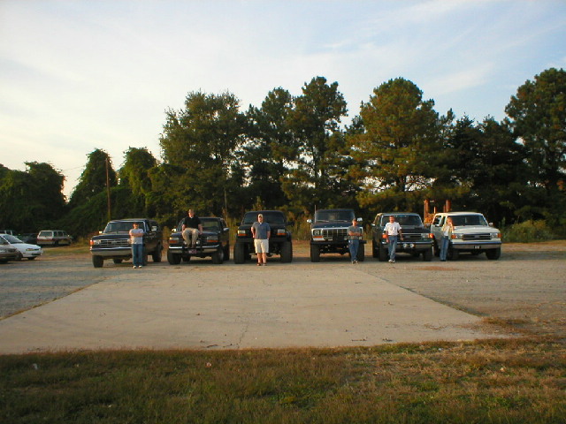 Six Ford Broncos and pickups parked in a gravel lot at dusk with owners standing between them