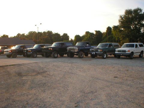 1989 Ford Bronco - A row of Ford Broncos, including a white one, a green one, and several black ones, are parked in a gravel lot.