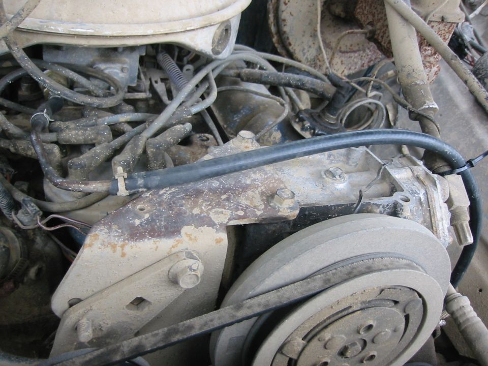 Dusty engine bay close-up showing A/C compressor pulley, belt, brackets, and spark plug wires