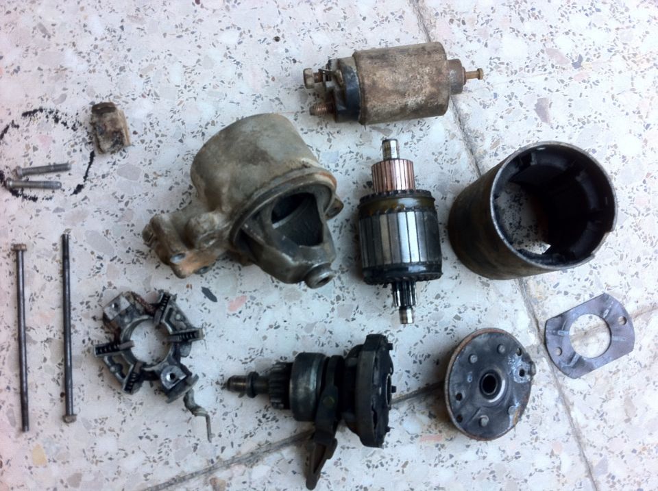 Starter motor components laid out on a tiled surface. Includes armature, field coil housing, solenoid, and brushes