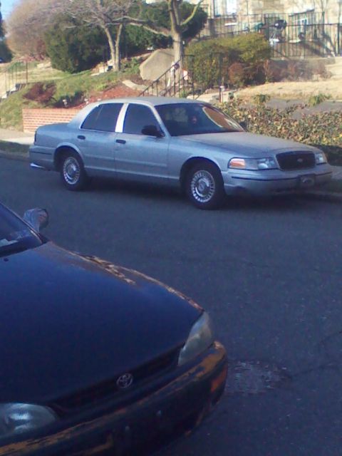 1999 Ford Crown Victoria - A silver 1999 Ford Crown Victoria is parked on the street, partially obscured by another vehicle.