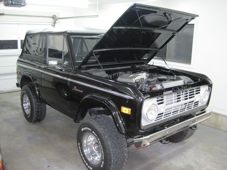Restored black early Ford Bronco in a garage with hood up showing a fuel-injected V8