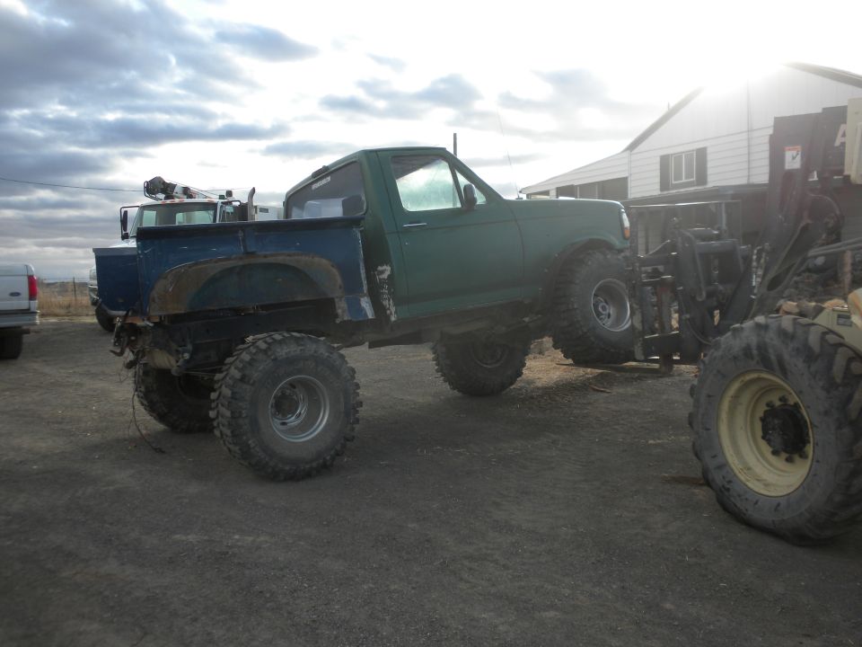 Green pickup truck with large tires being lifted by a forklift, another truck visible to the left