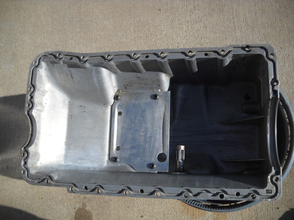Aluminum oil pan with a metal plate bolted on, resting on a concrete surface