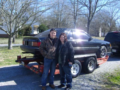 2007 Ford Mustang - A black 1987 Ford Mustang coupe is loaded onto a trailer, with two people standing beside it.