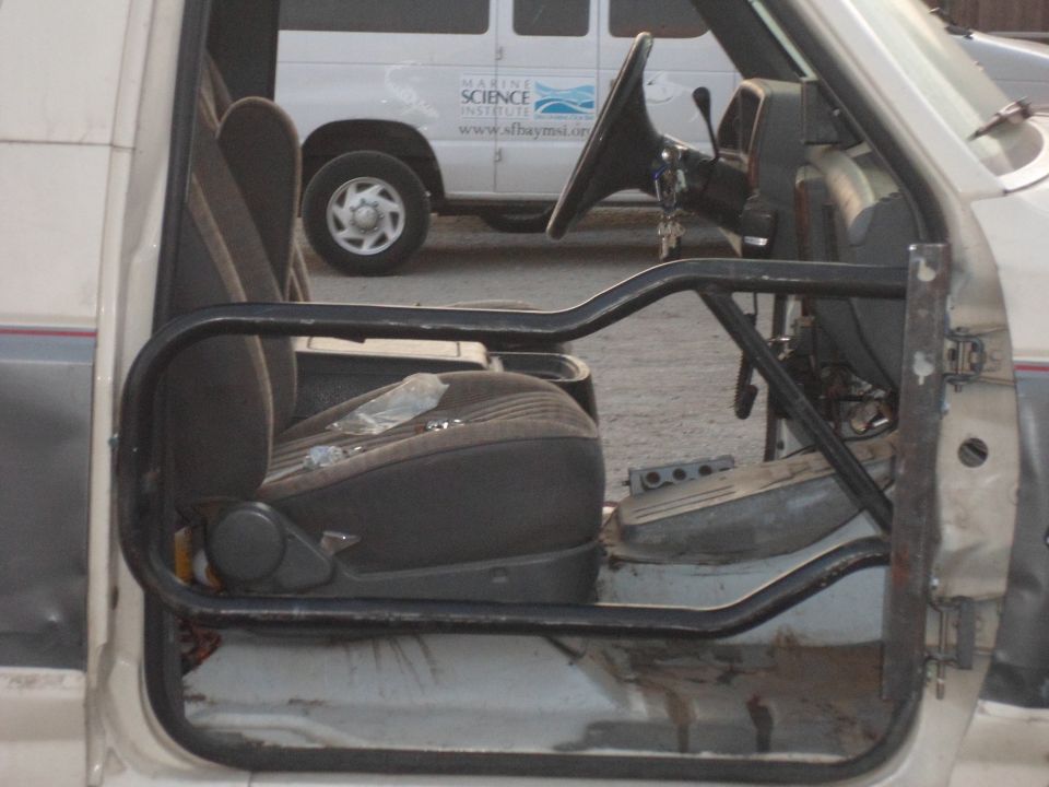 Interior shot of a vehicle with a roll cage, showing the driver's seat and steering wheel. A white van is visible in the background