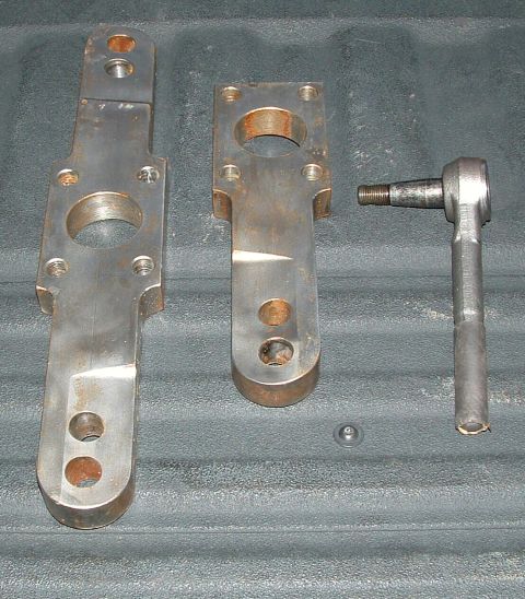 1979 Ford Bronco - Two bare-steel steering arms or pitman-style brackets laid out on a black plastic bedliner next to a tapered tie rod end with threaded stud…