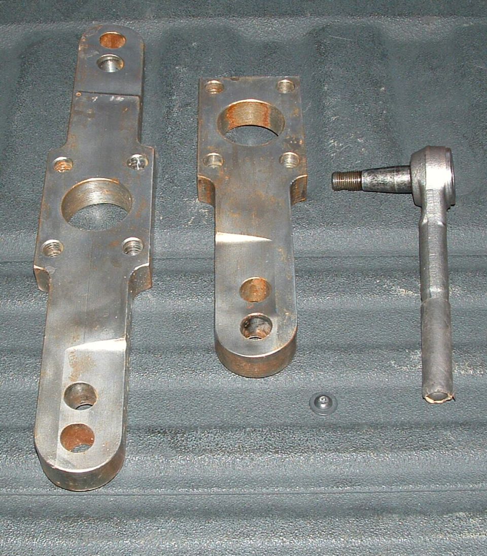 Two bare-steel steering arm brackets and a tie rod end laid on a black bedliner