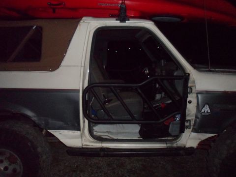 1991 Ford Bronco - Open driver's door of a 1991 Ford Bronco, revealing interior cage and a kayak on the roof rack.