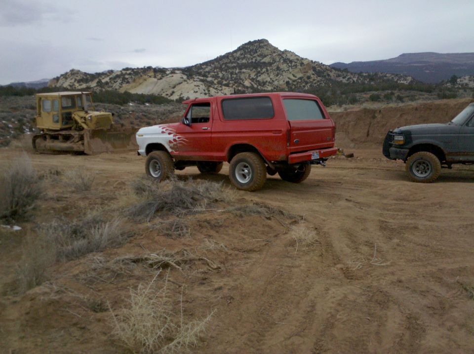 Red and white Ford Bronco with flame decals parked on dirt road, bulldozer and another SUV in background